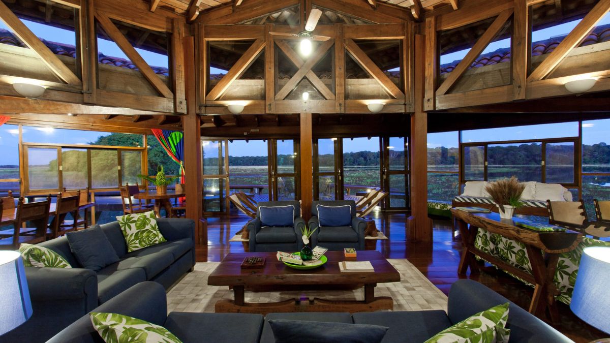 A spacious, elegantly designed living room with wooden beams and large windows overlooking a lush landscape. Comfortable sofas with green and white patterned cushions are arranged around a coffee table. Warm lighting creates a cozy atmosphere, complemented by decorative plants and artwork.