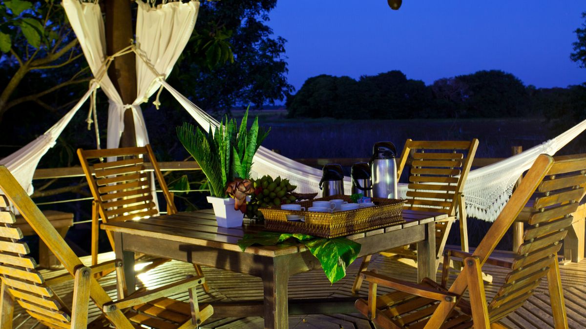 A cozy wooden patio at night, featuring a dining table surrounded by slatted chairs. A potted plant and a tray with snacks are on the table, flanked by two hammocks draping in the background. Soft lighting creates a warm atmosphere amidst lush greenery.