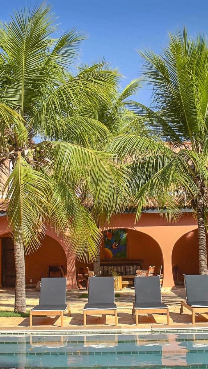 Lounge chairs by a swimming pool surrounded by tall palm trees and a warm-colored building. The sky is clear and blue, enhancing the tropical atmosphere.