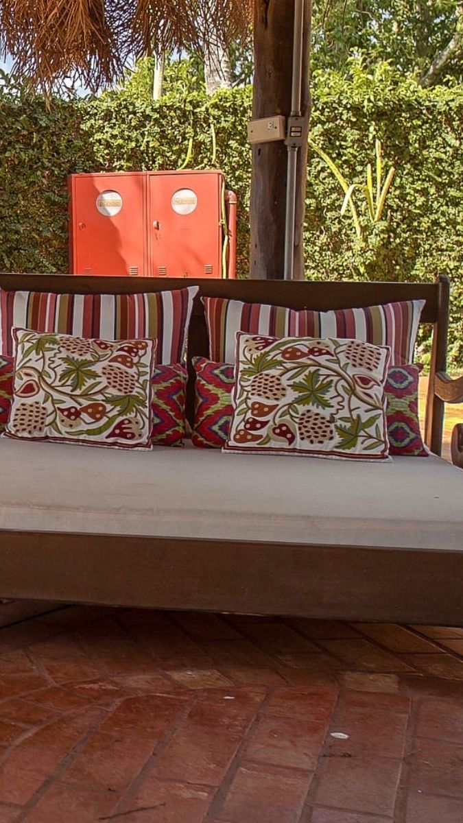 A wooden daybed adorned with patterned cushions sits under a thatched shelter, surrounded by lush greenery. Two red cabinets are visible in the background.
