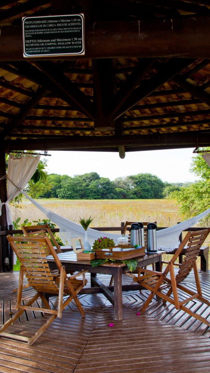 A rustic wooden deck features a round table set with coffee pots and decorative items. Hammocks are visible nearby, with lush greenery in the background. A sign detailing depth regulations is mounted on a wooden post.