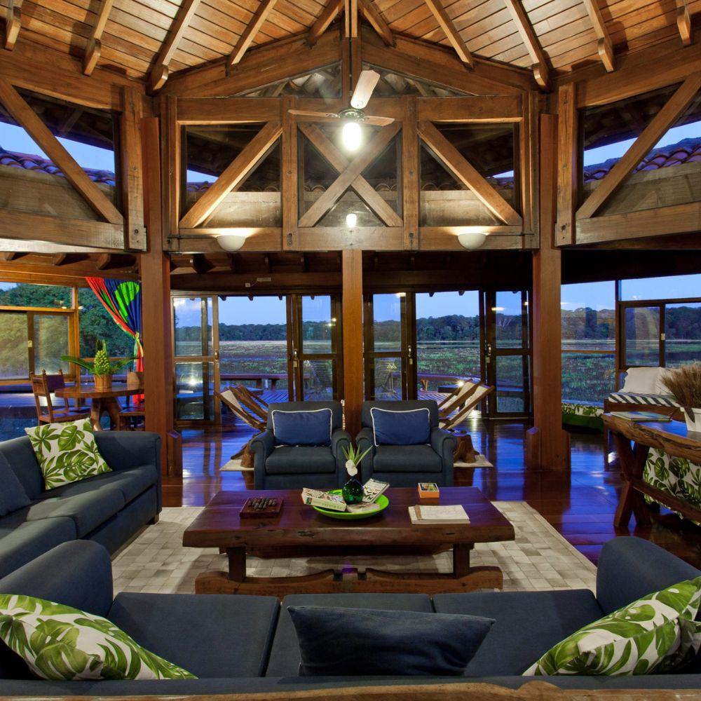 A spacious, rustic living room with high wooden ceilings and large glass doors opening to a scenic view. Two blue sofas are decorated with green cushions, and a wooden coffee table sits in the centre. Natural light fills the room, highlighting wooden beams and a cozy atmosphere.