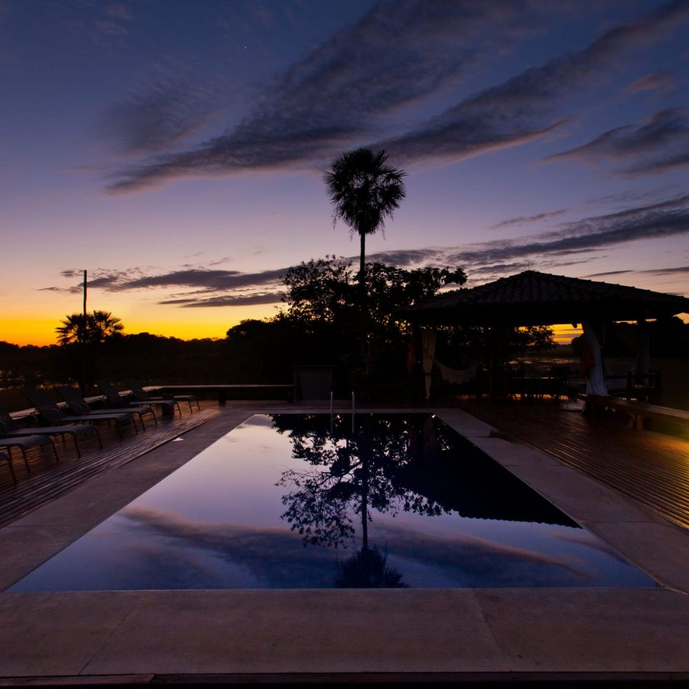 A serene sunset scene featuring a tranquil swimming pool reflecting the colorful sky. Silhouetted palm trees and a gazebo are visible in the background, surrounded by lush greenery.