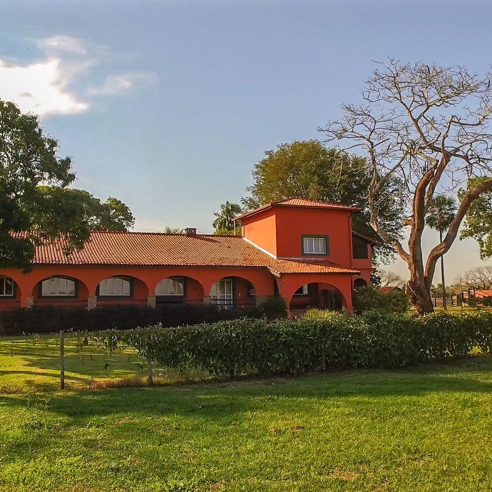 A vibrant red house with a tiled roof is surrounded by green grass and trees. The exterior features arched windows and a spacious porch, set against a clear sky.