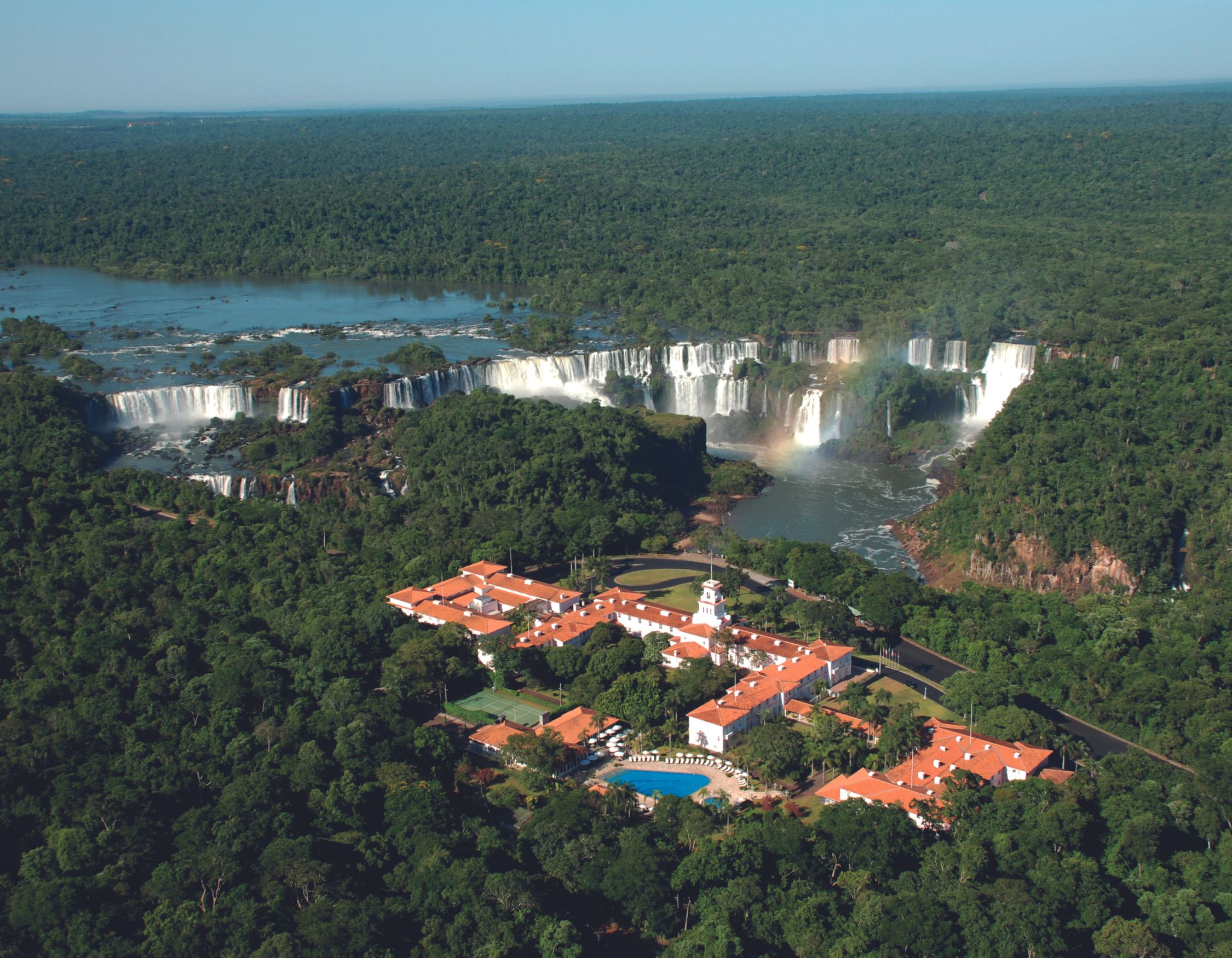 Aerial view of a large hotel surrounded by lush greenery, with a series of waterfalls in the background cascading into a river. The hotel features red rooftops and a swimming pool, set against a vibrant natural landscape.