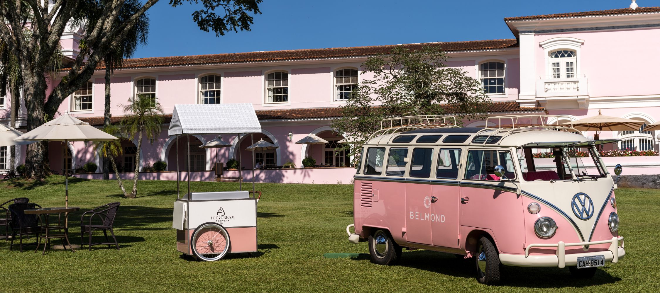 A vintage Volkswagen van in pastel pink parked on a grassy lawn, surrounded by trees and a large pink building in the background. A white parasol and a small seating area with chairs are visible nearby.