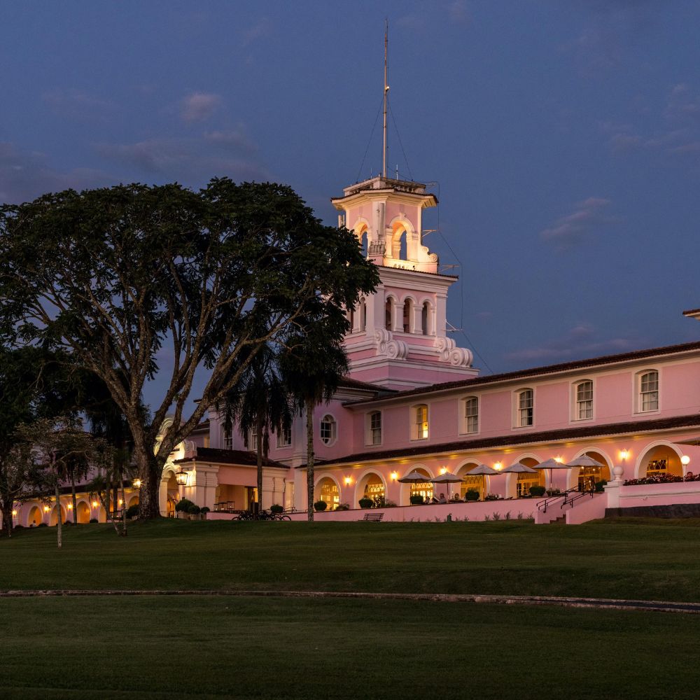 A historic pink building with a tall clock tower, illuminated by soft lights at dusk, set against a backdrop of trees and a grassy lawn.