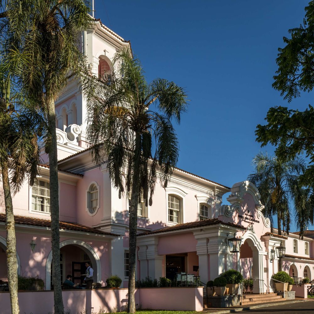 A charming pink building with a clock tower, surrounded by palm trees and lush greenery under a clear blue sky. The architecture features decorative elements and large arched windows.