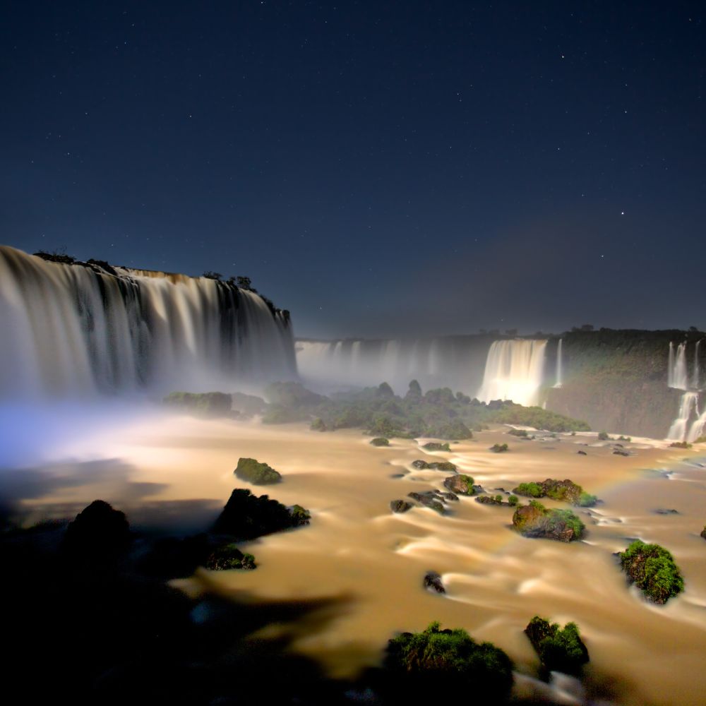 A breathtaking view of a waterfall at night, illuminated by stars. The cascading water creates a mist that gently envelops the rocky shore, while soft light reflects off the smooth surface of the water below.