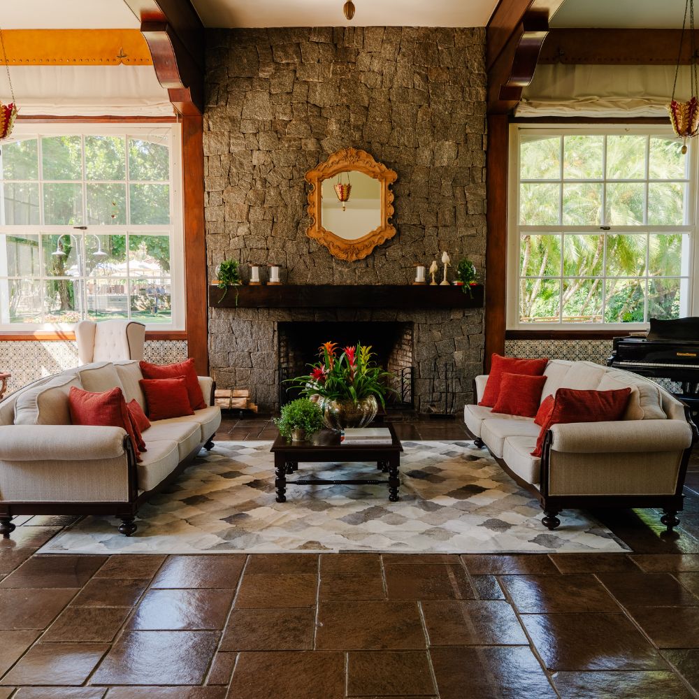 A cozy living room featuring two light-colored sofas with red cushions facing a coffee table. A stone fireplace is centrally located, adorned with a vintage mirror above it. Large windows allow natural light in, and lush greenery is visible outside. A patterned rug covers the floor.