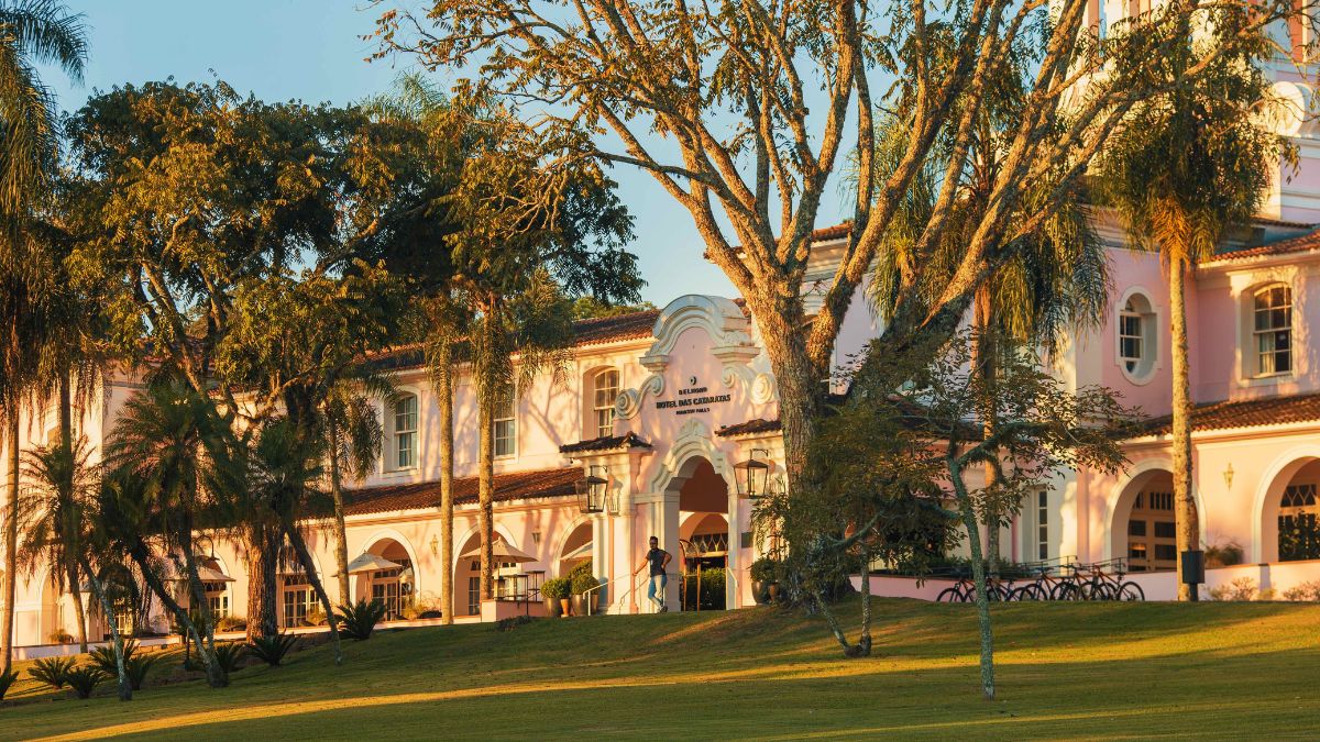 Sunlight casts a warm glow on a charming building with white and pastel pink architecture, surrounded by lush greenery and tall trees. The entrance features an archway, and bicycles are parked nearby on a grassy lawn.