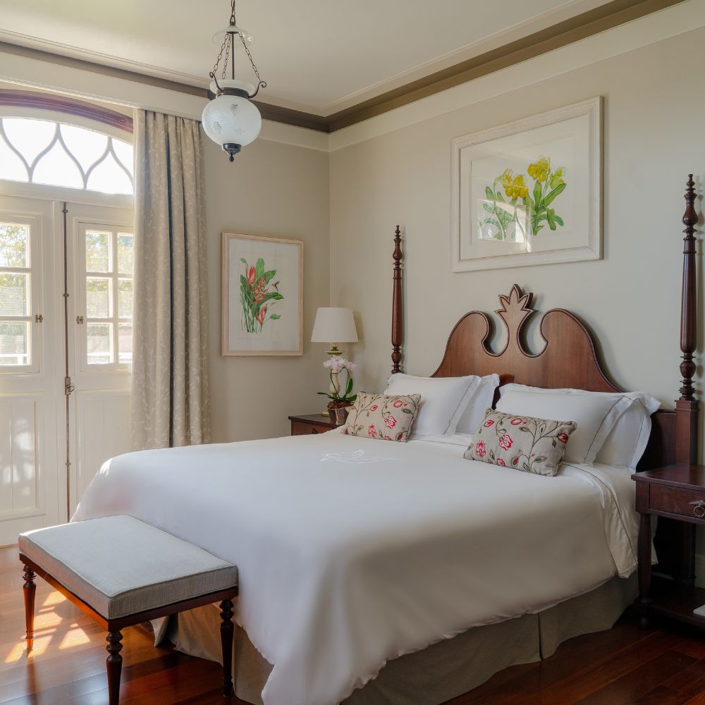 A cozy bedroom featuring a large bed with white linens and decorative pillows. A wooden bench sits at the foot of the bed. The room includes framed botanical artwork on the walls, light gray curtains, and a vintage lamp on a bedside table. An elegant window with ornamental arches allows natural light to fill the space.
