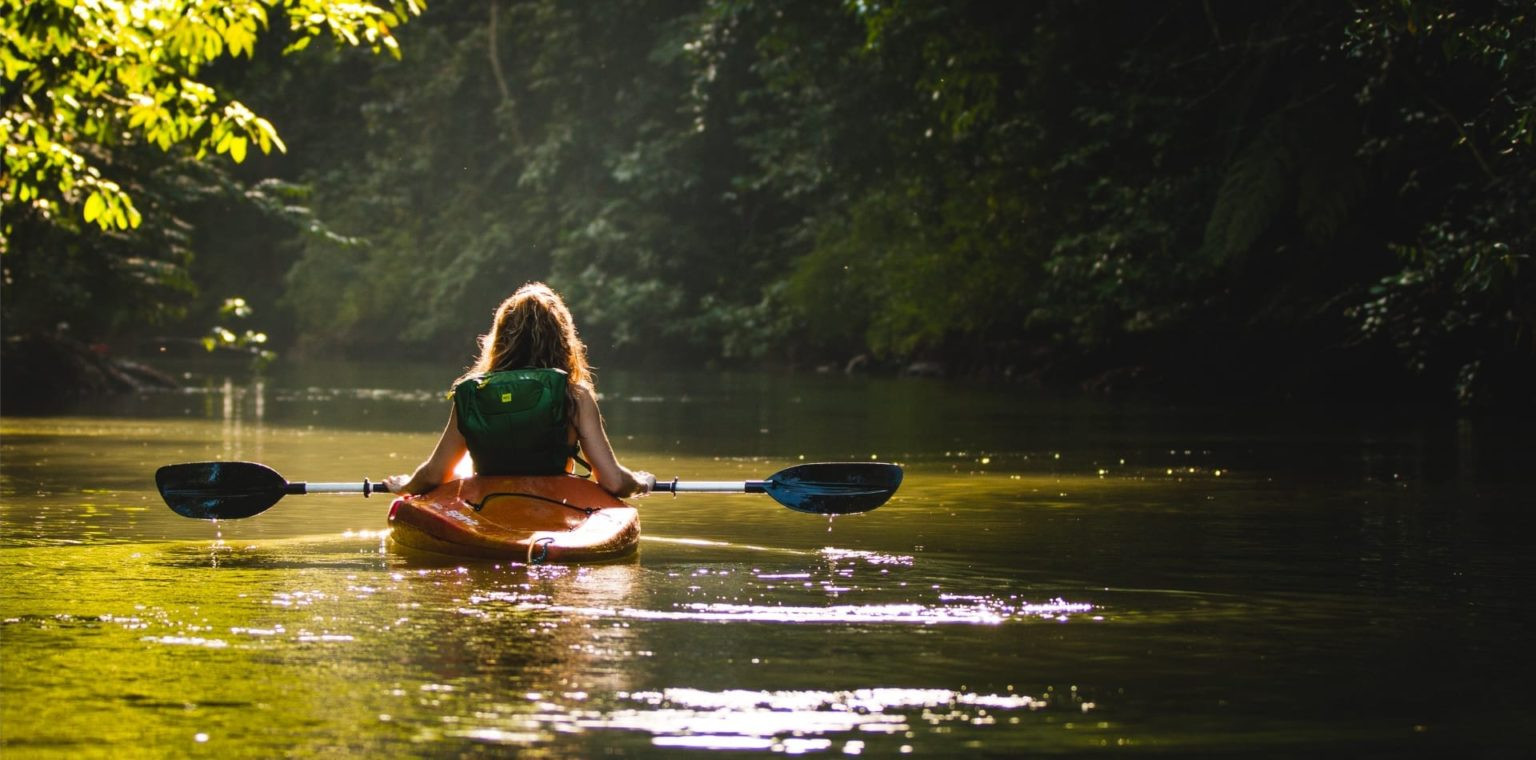 A person with long hair is kayaking down a serene, tree-lined river. Sunlight filters through the leaves, creating a calm and tranquil atmosphere. The kayak is orange, and the paddles are blue, reflecting in the water.