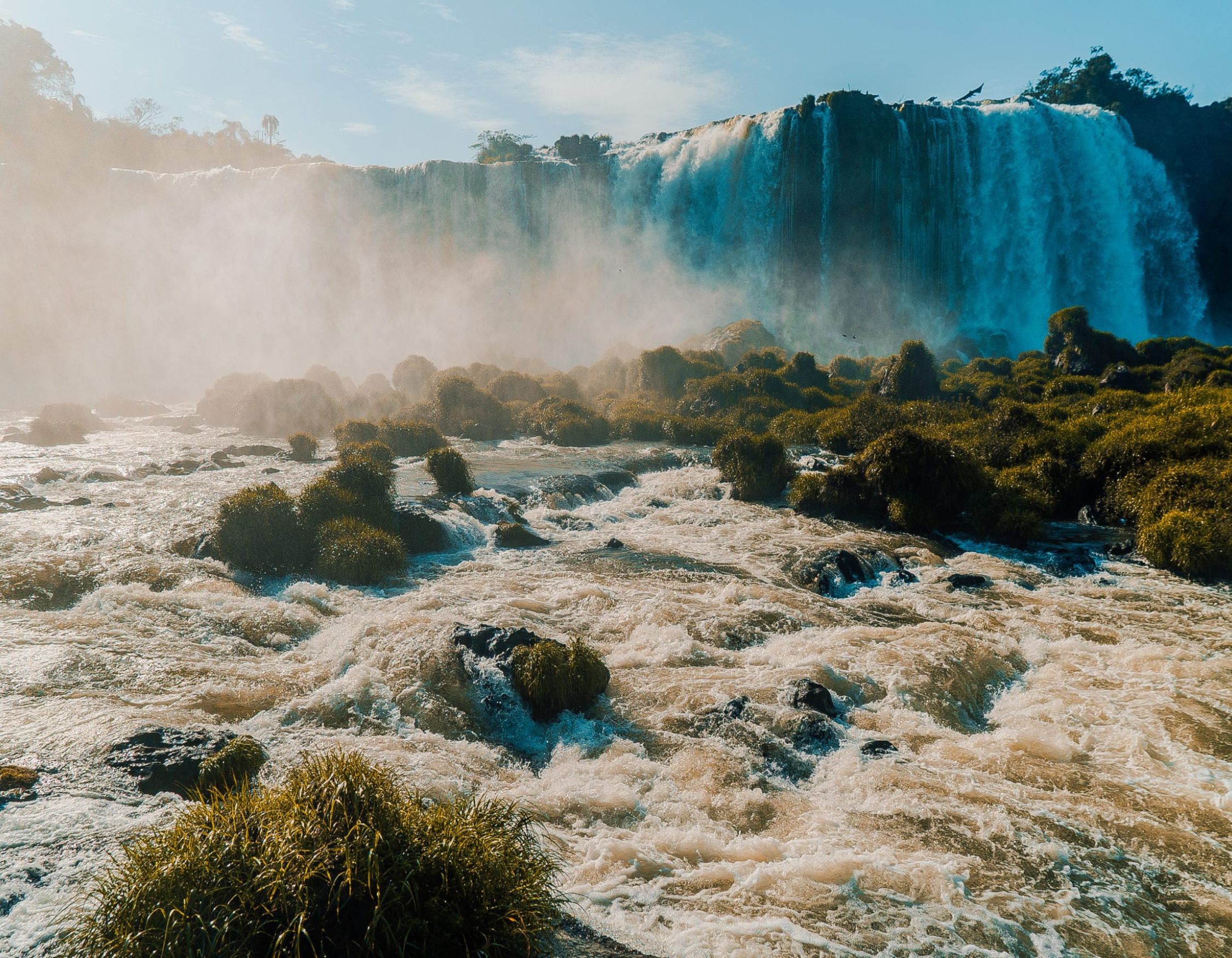 A stunning waterfall cascading down rocky cliffs, surrounded by mist and lush greenery. The river flows strongly at the foreground, with patches of grass and rocks visible. Sunlight filters through the mist, creating a serene atmosphere.