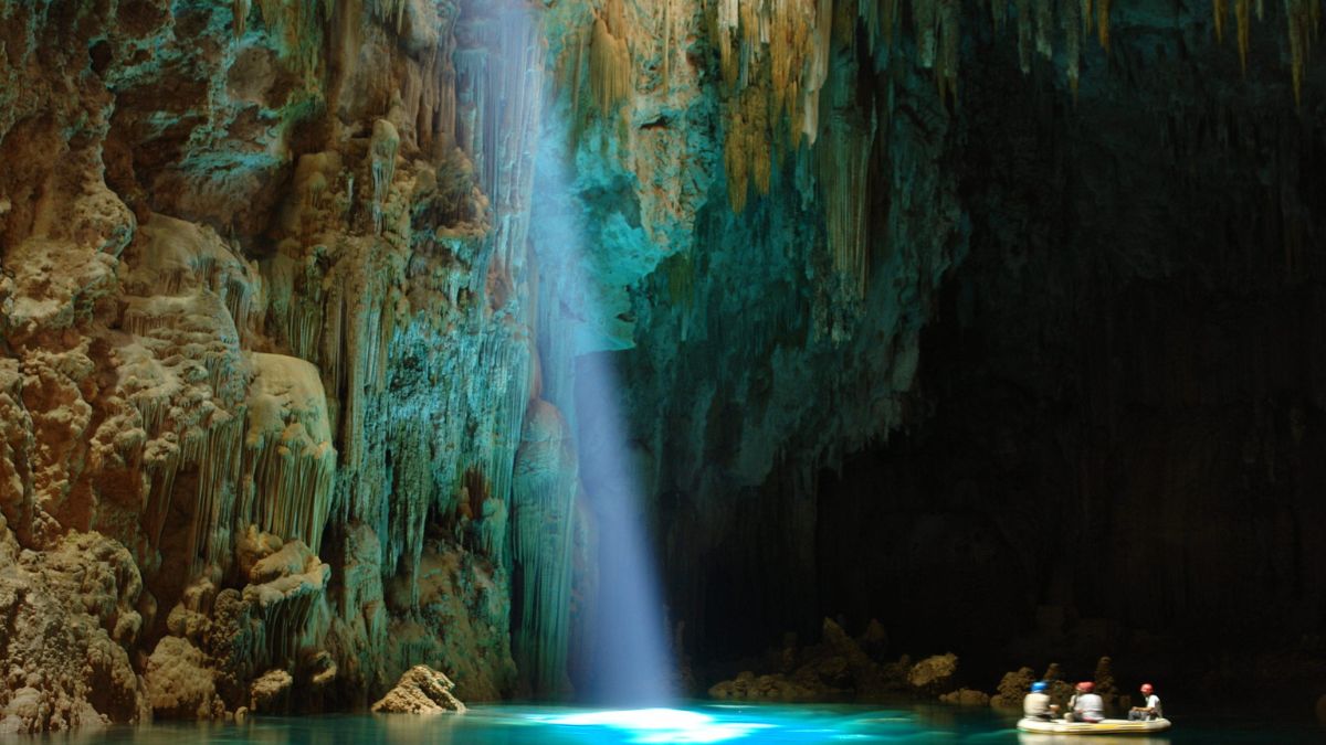 A stunning cave interior with towering stalactites and a glowing blue underground lake. A beam of light shines down from above, illuminating the water and highlighting the natural formations. A small boat with four people is visible in the foreground, adding a sense of scale to the scene.