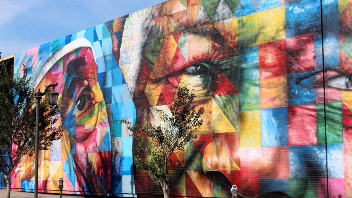 Vibrant mural featuring abstract faces with geometric shapes in bold colours. The artwork showcases intricate details and a striking mix of hues, creating a lively visual impact against a blue sky.