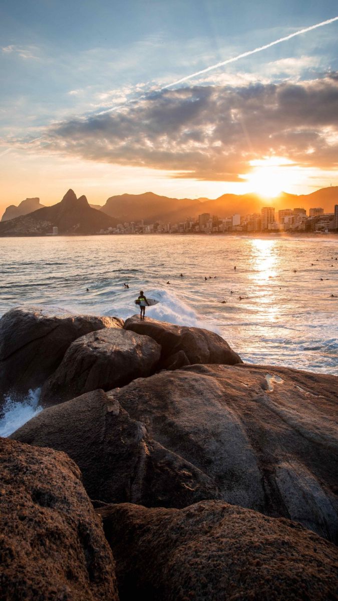 A lone surfer stands on large rocks by the shore, watching the sunset over the ocean. Waves crash against the rocks, and the skyline of a coastal city is visible in the background, framed by mountains under a partly cloudy sky.