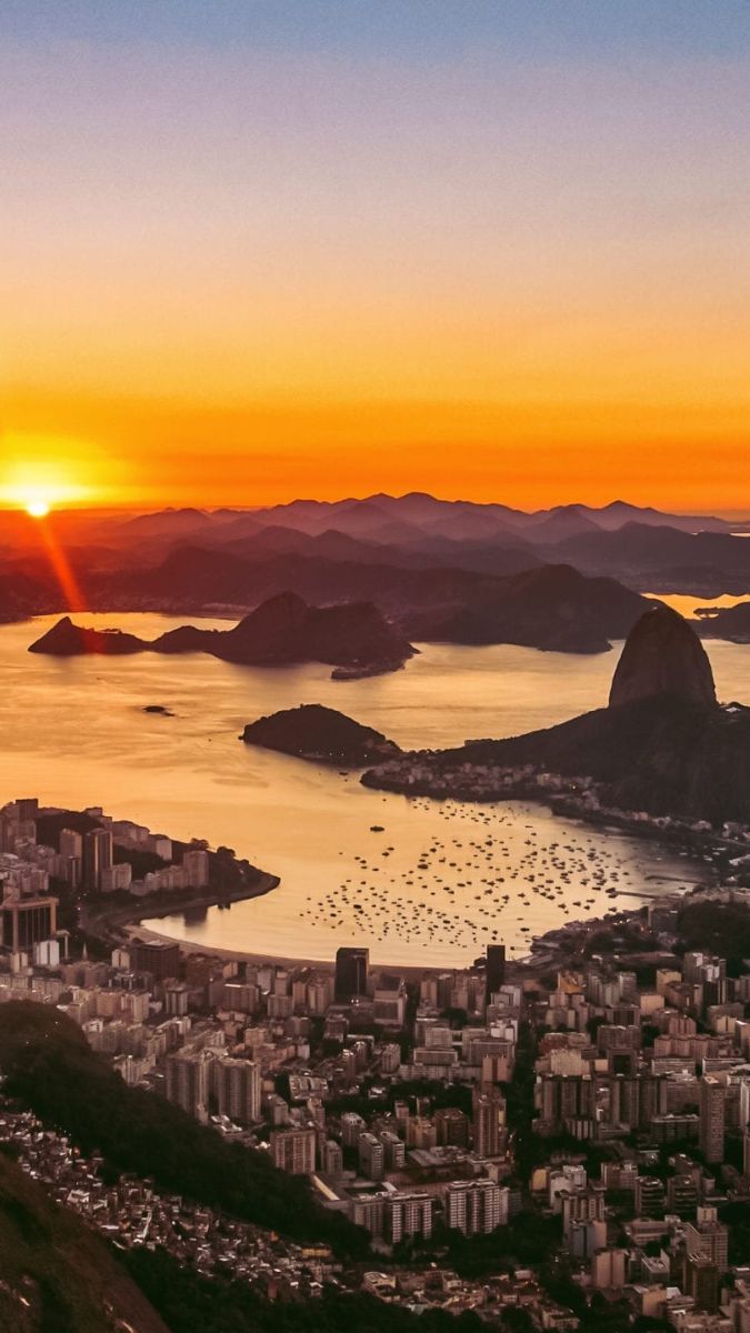 Breathtaking sunset over Rio de Janeiro, with vibrant orange and yellow hues lighting up the sky. The silhouette of Sugarloaf Mountain stands prominently against the horizon, while the bay reflects the warm colors. The cityscape is visible in the foreground, surrounded by mountains in the distance.