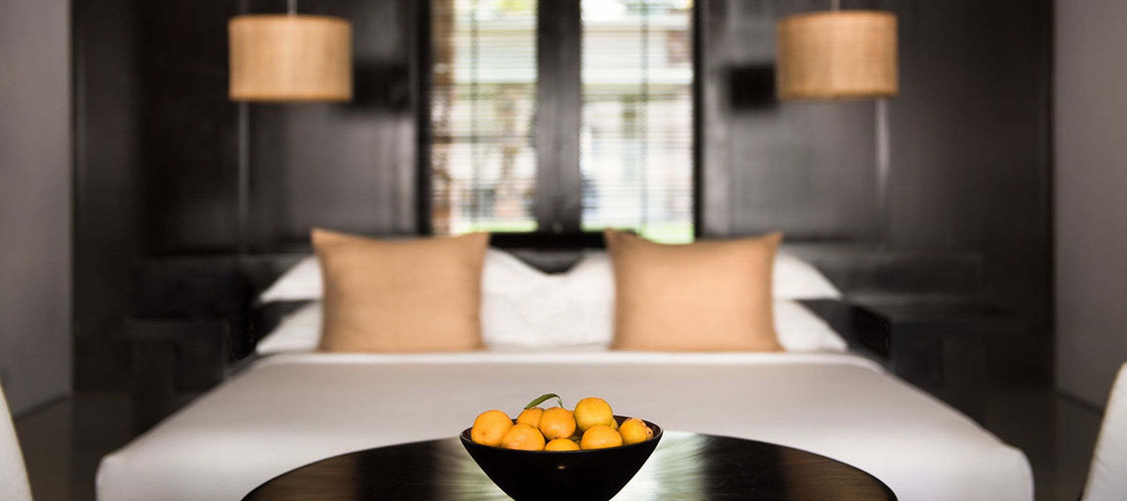 A cozy bedroom featuring a neatly made bed with white linens and two brown decorative pillows. In the foreground, there's a black bowl filled with small yellow fruits, set on a circular table. Soft lighting is provided by pendant lamps hanging from the ceiling.