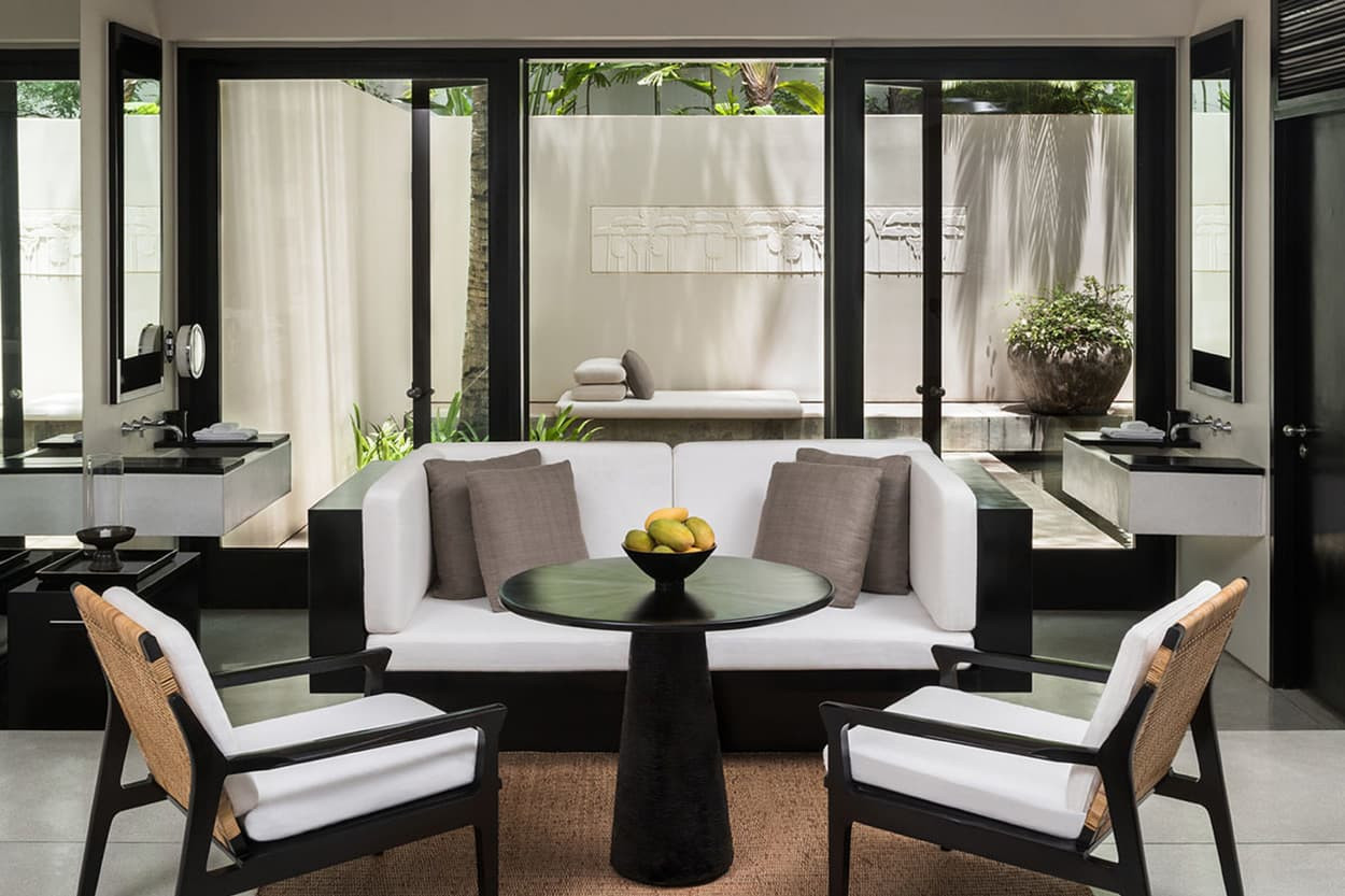 A modern, stylish living room featuring a circular black table surrounded by white sofas with gray cushions and two black chairs. Large windows offer a view of a serene outdoor area with greenery.