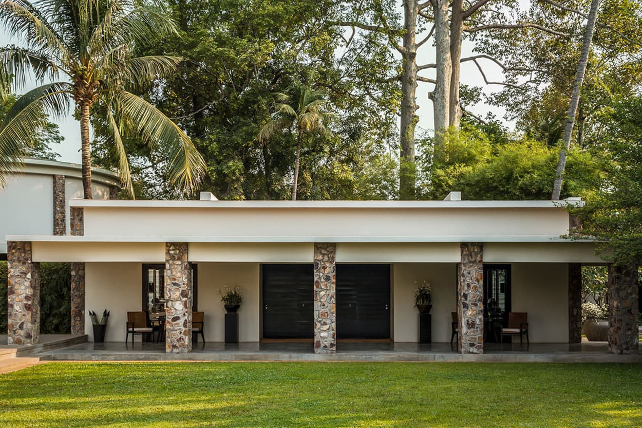 A modern single-story house featuring a flat roof and stone accents, surrounded by lush greenery and palm trees. The facade has large wooden doors and potted plants on either side of the entrance.
