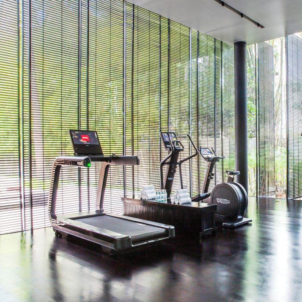 A modern gym space featuring a treadmill, elliptical trainer, and stationary bike. The room is surrounded by large windows with slender blinds, allowing natural light to filter through. Bottled water is neatly arranged on a bench.
