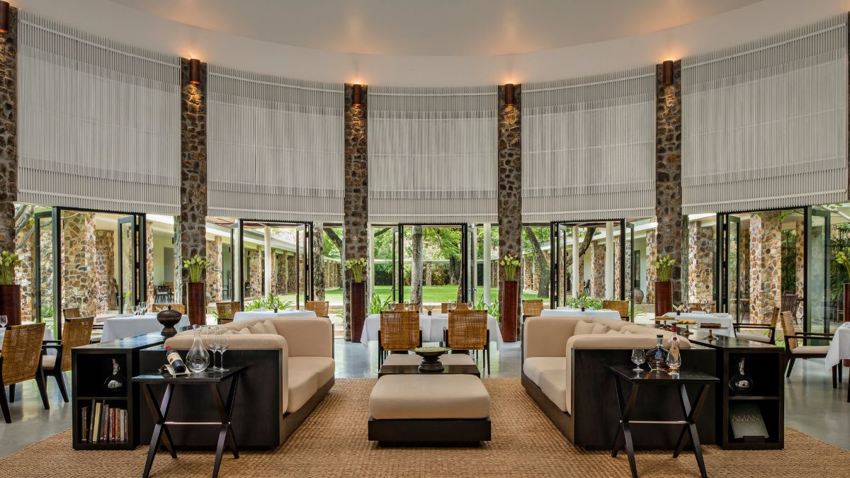 Modern restaurant interior featuring large windows, stone walls, and stylish furniture. Comfortable sofas are placed around a central coffee table on a woven rug. Bright and open atmosphere with greenery visible outside.