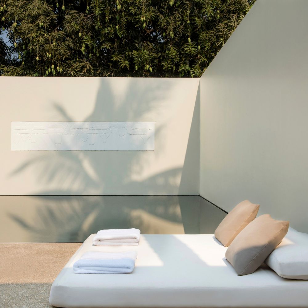 A serene outdoor space features a minimalist sunbed with beige cushions by a reflective pool, surrounded by lush green foliage and a textured wall.