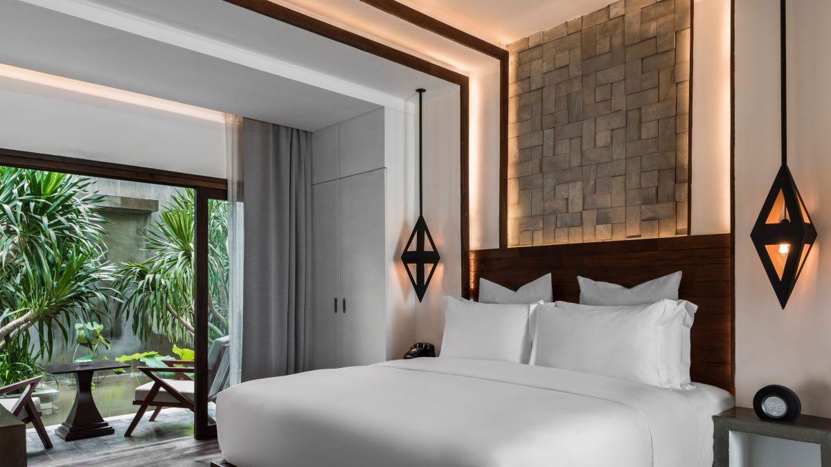 A modern bedroom featuring a large, neatly made bed with white linens. The stylish room has wooden accents, geometric wall decor, and warm lighting. Large glass doors lead to a lush, private outdoor area with greenery. Two elegant pendant lights hang beside the bed.