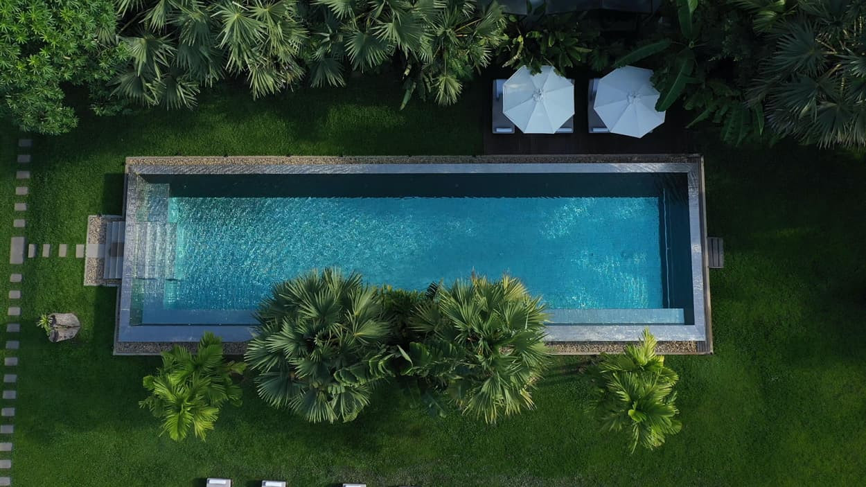 Aerial view of a rectangular swimming pool surrounded by lush green foliage and palm trees. Two white lounge chairs with umbrellas are visible beside the pool. Soft stone pathways lead through the grass.