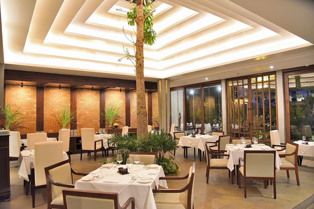 A modern restaurant interior featuring elegant white tablecloths on dining tables, surrounded by comfortable chairs. The ceiling has a unique, wavy design with soft lighting, and there are greenery and a small tree inside, enhancing the ambiance. Large glass windows reveal a garden view.