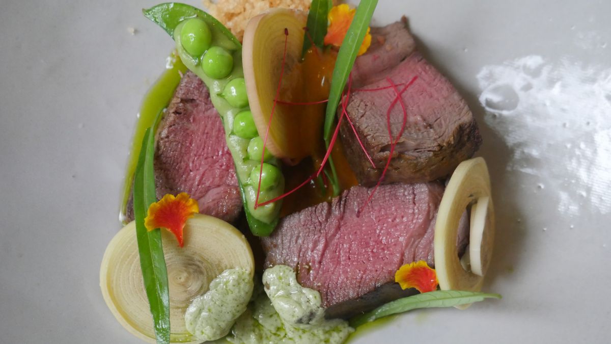 Sliced medium-rare beef garnished with green peas, spring onions, and vibrant edible flowers, drizzled with a green sauce and accompanied by crispy elements.