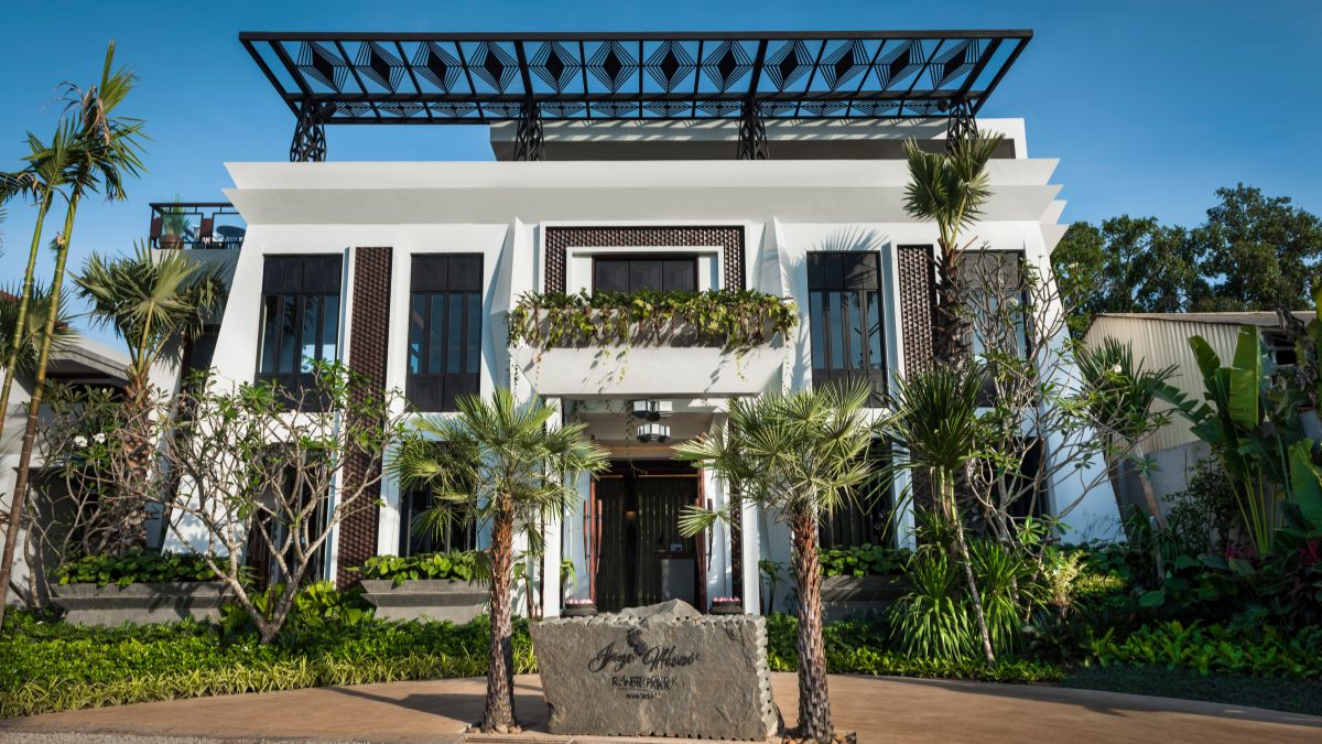 A modern, two-story building with a stylish façade featuring large windows and greenery. The entrance is framed by tropical plants and a decorative stone sign. Clear blue sky in the background.