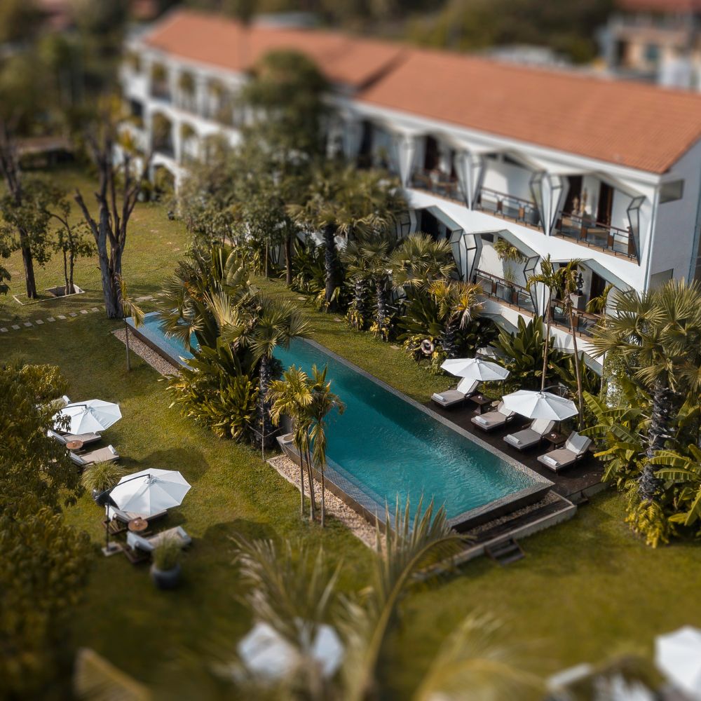 A lush green landscape features a narrow swimming pool surrounded by sun loungers and white umbrellas, set against a backdrop of a modern building with balconies. Palm trees and other tropical plants enhance the serene atmosphere.