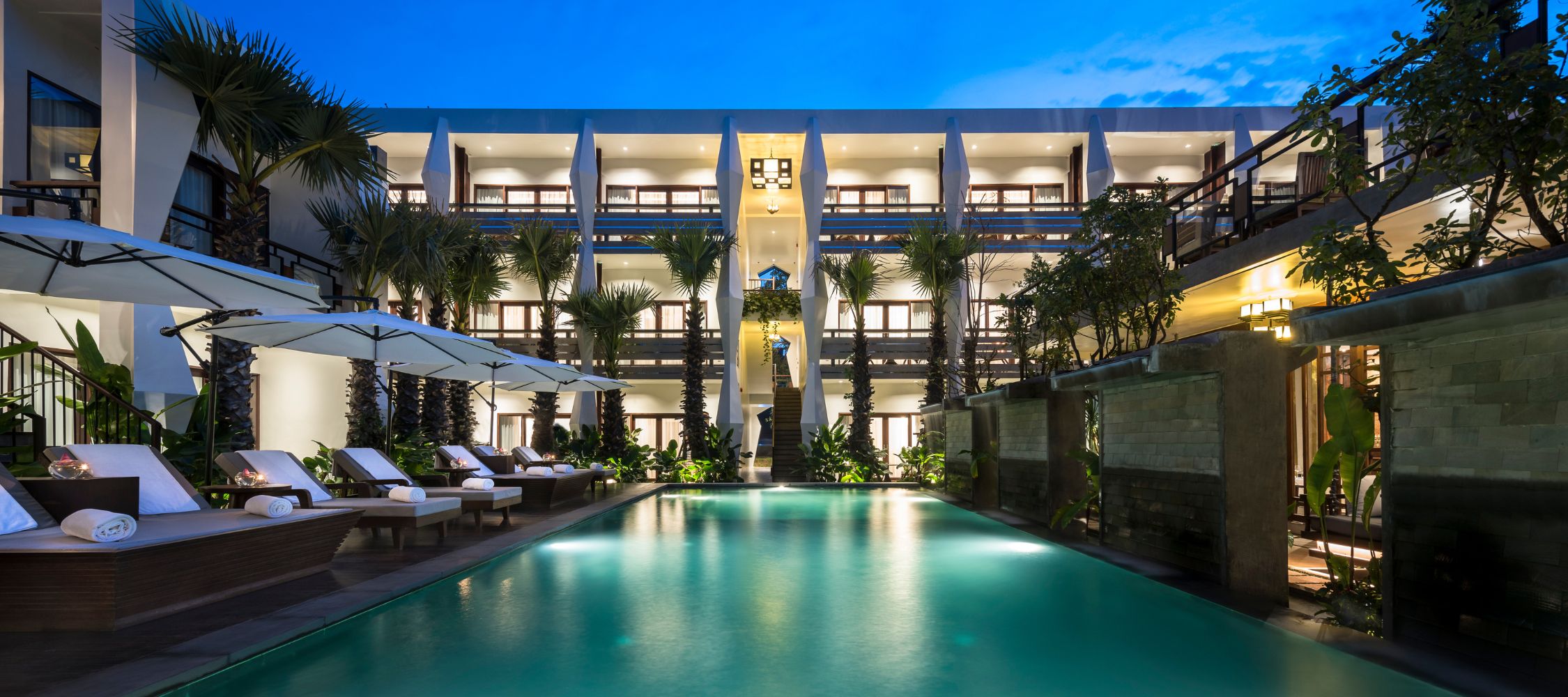 A serene hotel courtyard featuring an illuminated pool surrounded by lounge chairs and palm trees. The building's architecture showcases multiple balconies, set against a twilight sky.