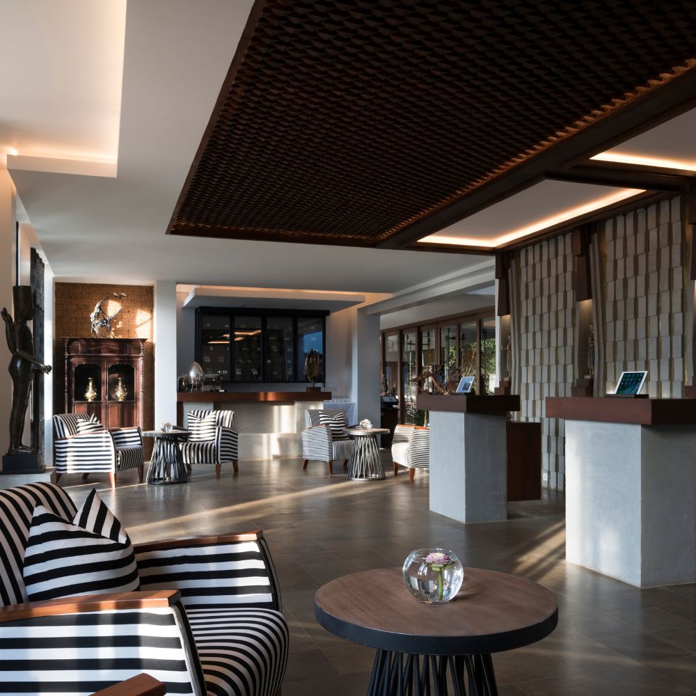 A modern hotel lobby featuring stylish striped chairs, a circular table, and sleek reception desks. Large windows allow natural light to fill the space, highlighting wooden and stone elements in the design.