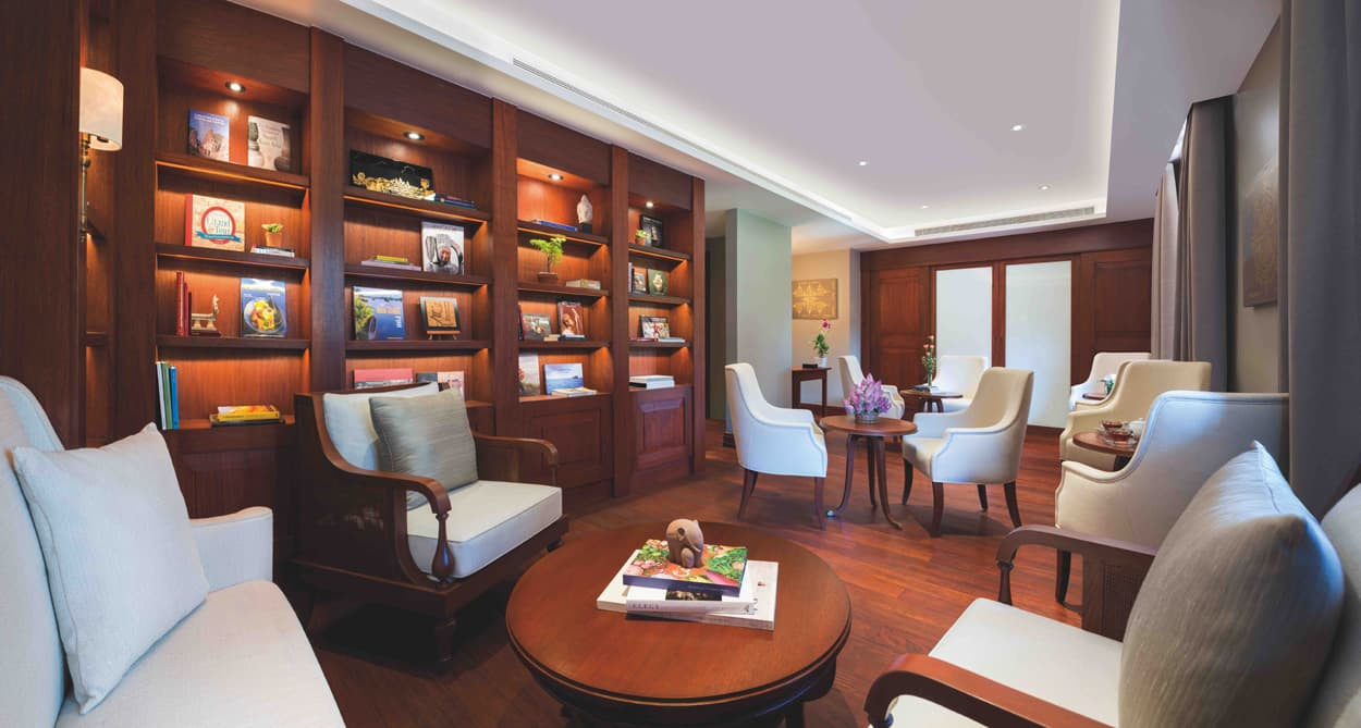 A cozy lounge area featuring comfortable armchairs and a wooden coffee table, surrounded by shelves filled with books and decorative items. Soft lighting highlights the warm wooden tones and inviting atmosphere.