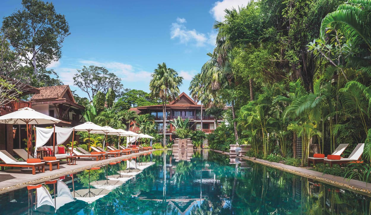 A luxurious resort featuring a serene pool surrounded by tropical palm trees and sun loungers, with a beautifully designed building in the background under a clear blue sky.