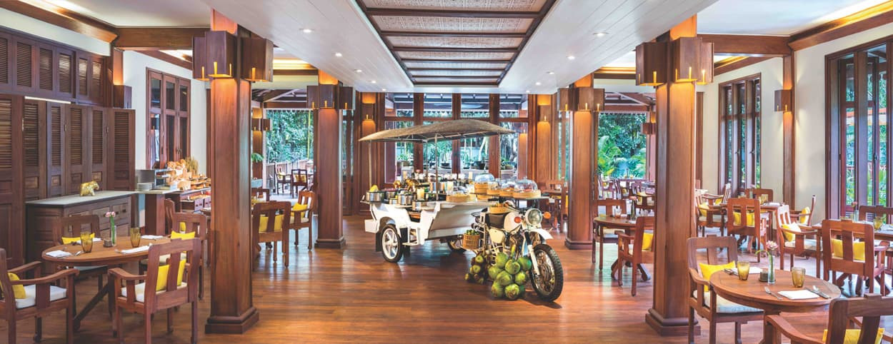 A spacious restaurant with wooden decor, featuring tables and chairs arranged throughout. In the center, a unique food cart on a motorcycle is displayed, surrounded by tropical greenery and large windows that allow natural light to fill the space.