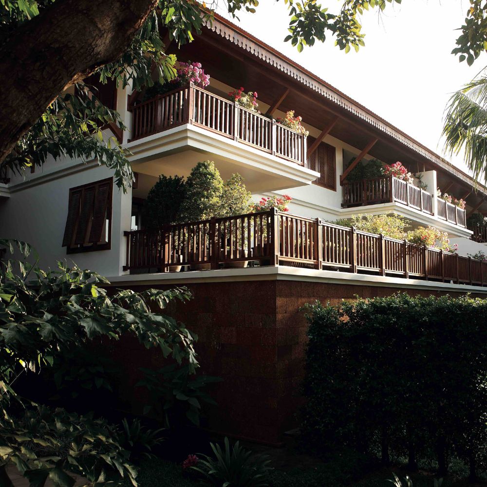 A modern, multi-storey building with wooden balconies adorned with potted plants, surrounded by lush greenery and tropical trees. The sunlight casts a warm glow on the structure, highlighting its architectural details.