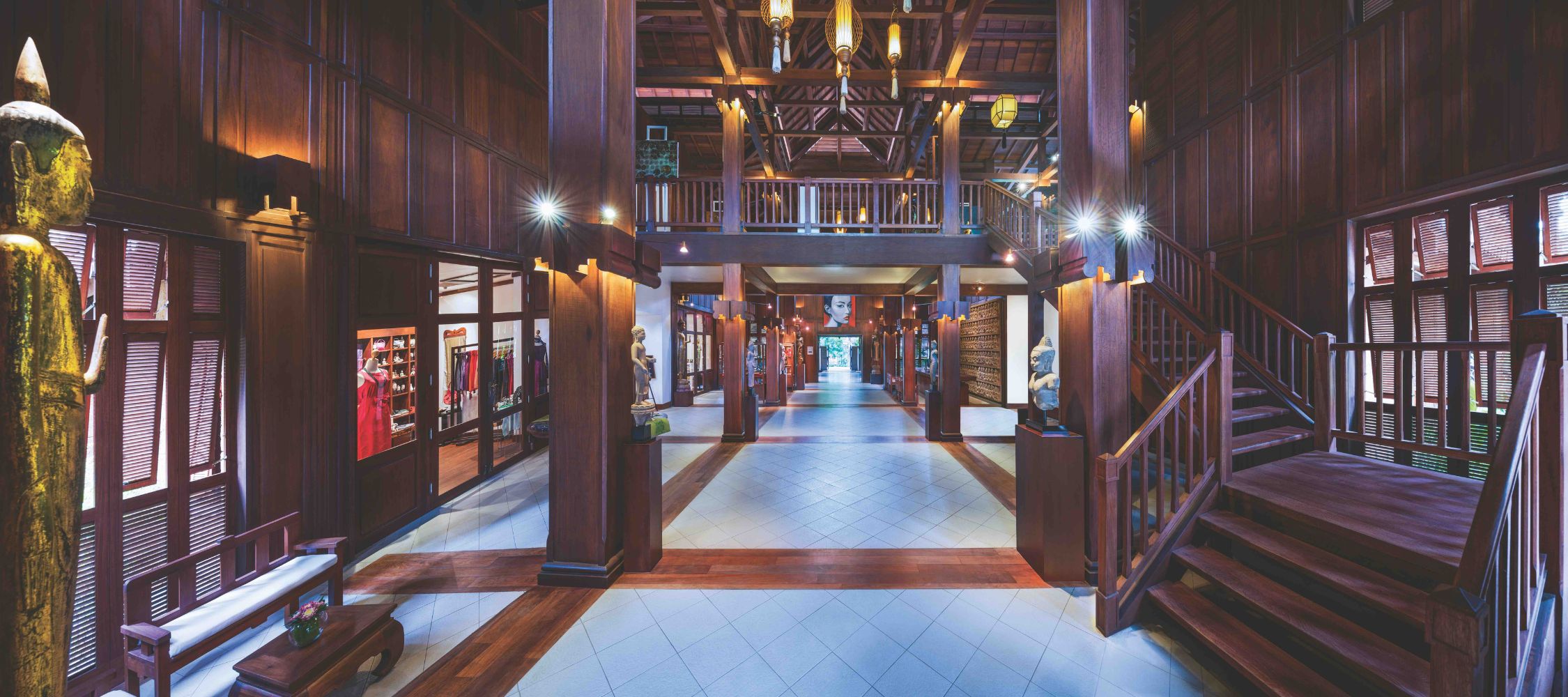 A spacious wooden interior showcasing a traditional architectural design, featuring multiple corridors leading to rooms, decorative wooden beams, and warm lighting. Statues and art pieces are displayed along the walls, enhancing the cultural ambiance.