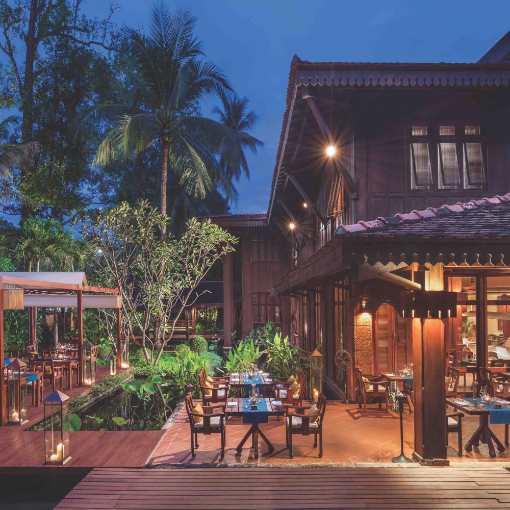 A beautifully lit outdoor dining area at a tropical restaurant, surrounded by lush greenery and palm trees, with wooden tables set for guests. The building features traditional architecture with a warm wooden façade. вечернее освещение создает уютную атмосферу.