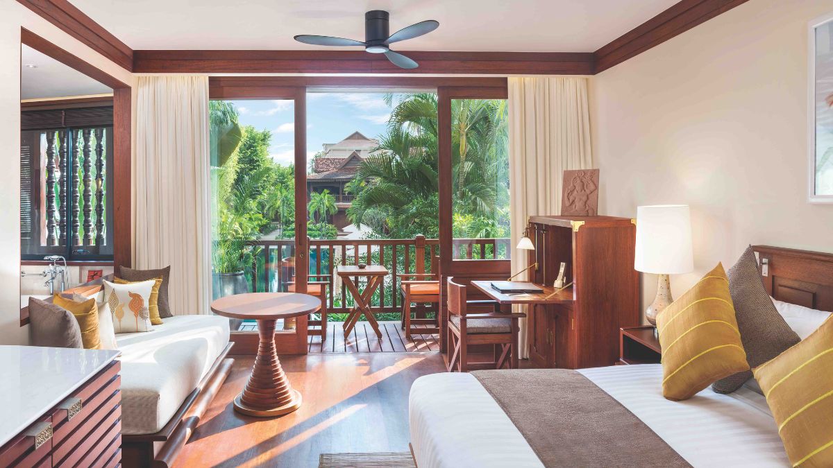 Bright, airy hotel room featuring a king-sized bed, a small table, and a seating area. Large windows showcase lush green tropical foliage outside. The decor is modern and warm, with wooden furnishings and soft, neutral colors.