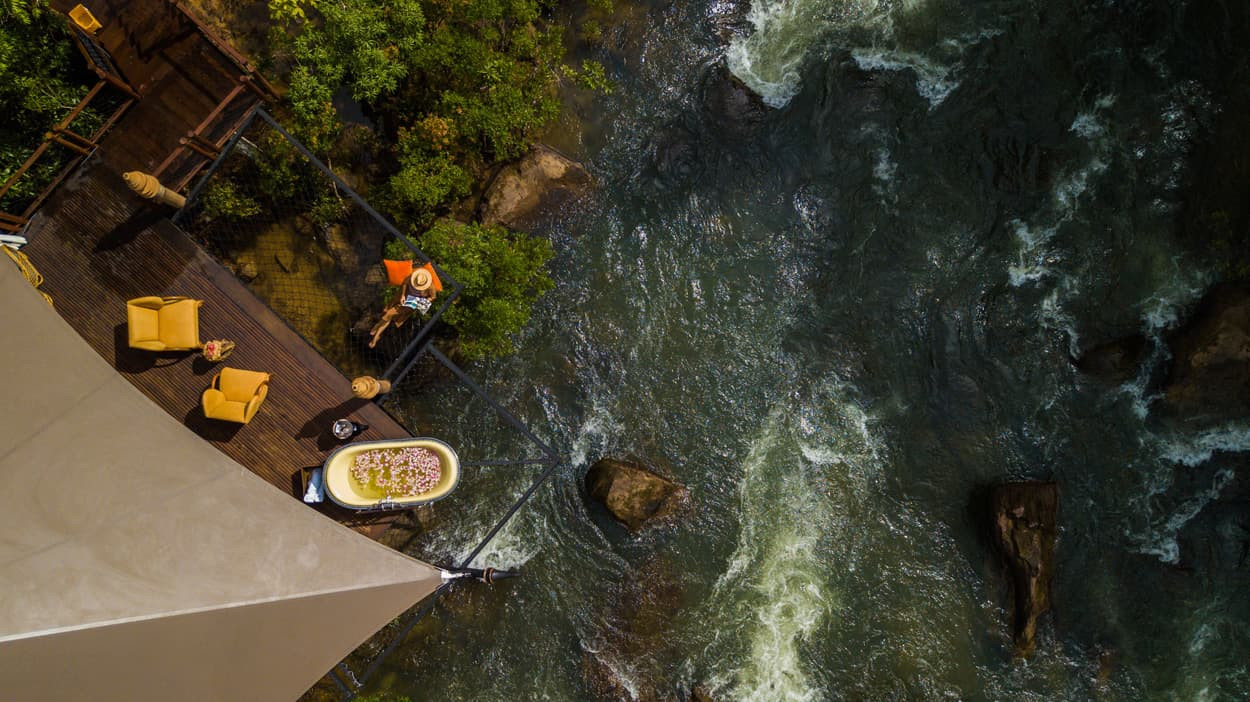 Aerial view of a riverside deck featuring two yellow chairs, a bathtub filled with flowers, and a person standing nearby. The flowing water and lush greenery surround the space, creating a serene atmosphere.