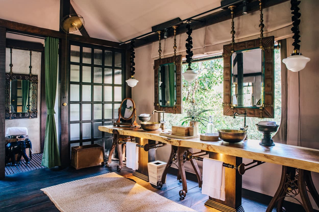 A spacious, elegantly designed bathroom featuring a long wooden vanity with two sinks, ornate mirrors, and unique lighting fixtures. Natural light streams in through large windows, highlighting the greenery outside. The decor combines rustic and modern elements, creating a serene atmosphere. A woven rug lies on the floor, enhancing the overall aesthetic.