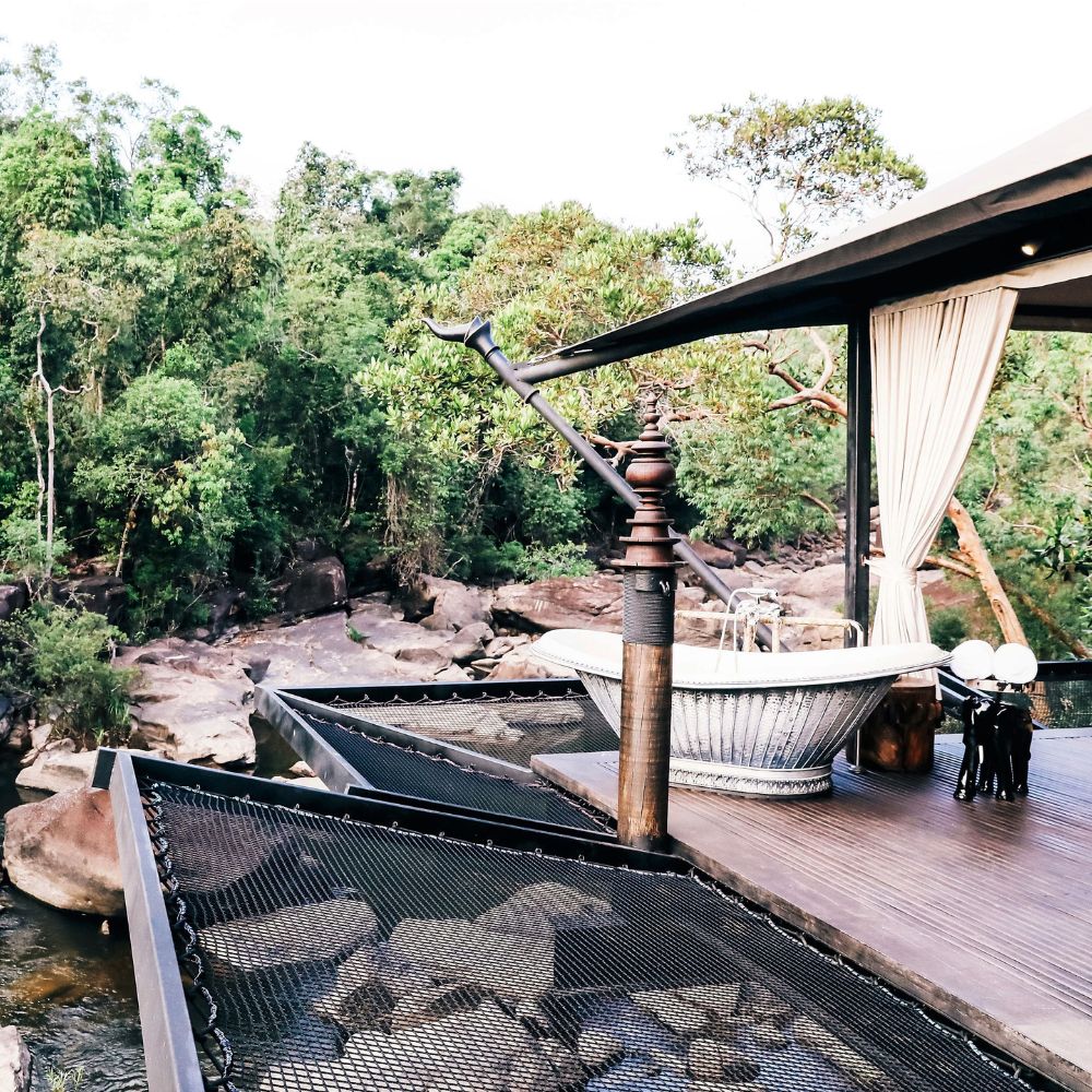 A luxurious outdoor bathing area overlooks a rocky riverbank, surrounded by lush greenery. A stylish bathtub sits on a wooden deck, with elegant drapes that create a serene atmosphere.