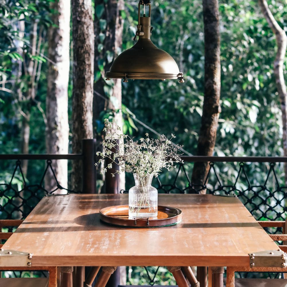 A wooden table in a forest setting, featuring a glass vase with dried flowers and a round wooden tray, illuminated by a brass pendant light hanging above. Lush greenery surrounds the scene.