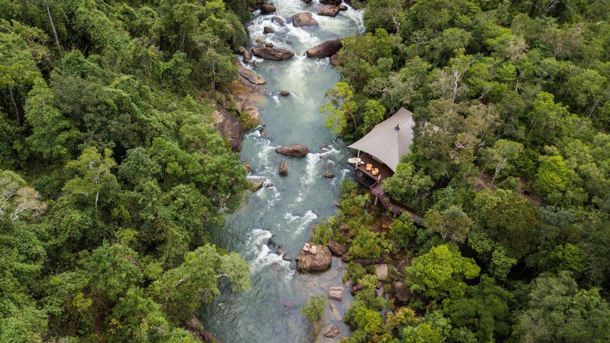 Aerial view of a tented structure beside a flowing river, surrounded by dense green foliage and rocky terrain. The serene landscape features clear water and large stones scattered throughout the river.