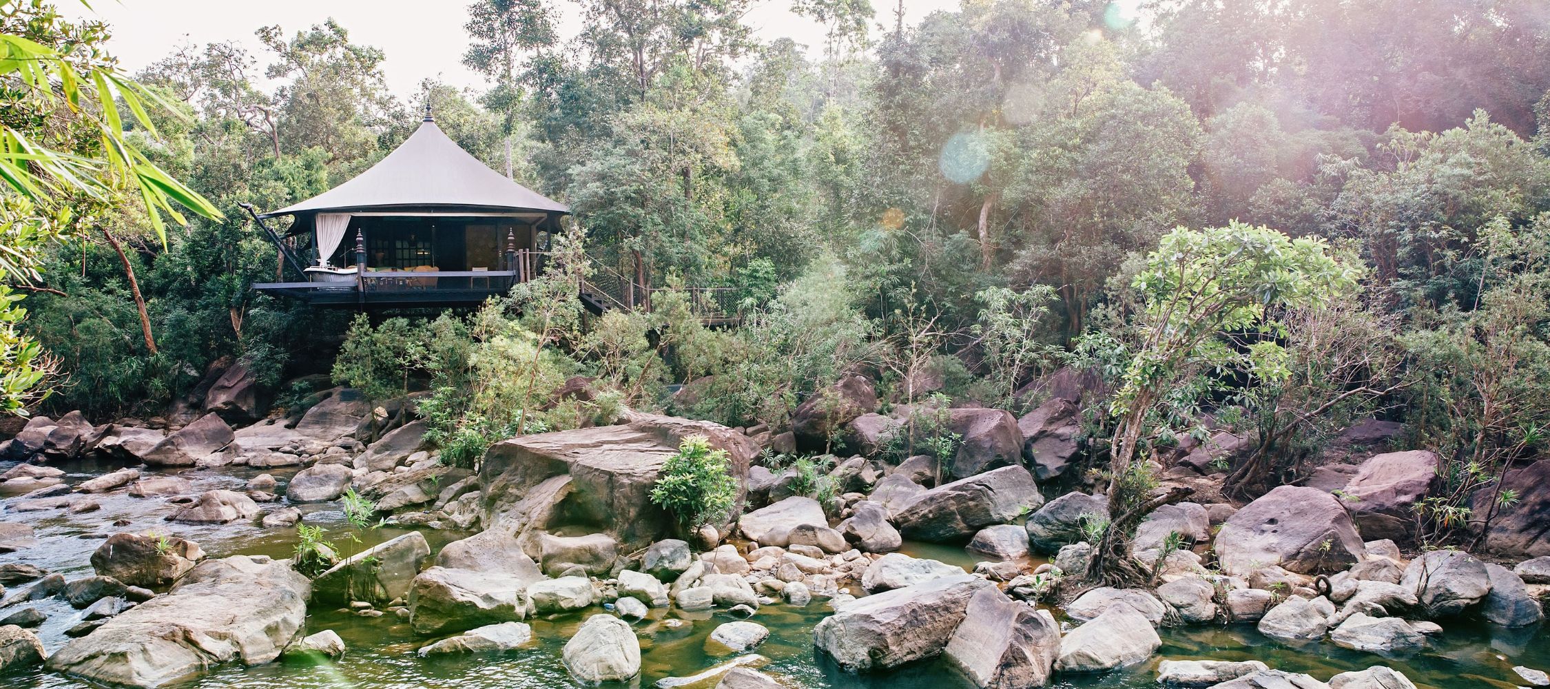 A modern tented cabin perched above a rocky riverbank, surrounded by dense greenery and trees. Sunlight filters through the foliage, creating a serene atmosphere.