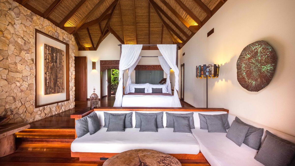 A spacious, stylish bedroom featuring a four-poster bed draped with white fabric, surrounded by natural stone walls and wooden flooring. A large, cushioned seating area with grey pillows is in the foreground, adding to the luxurious ambiance. Elegant lighting enhances the warm, inviting atmosphere.